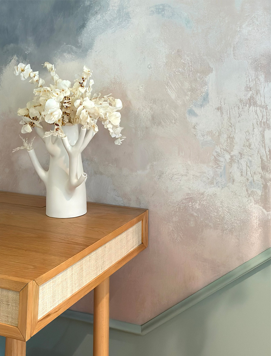 Styled oak and raffia desk vignette with a sculptural white branch vase and dried white stems against a soft blue and blush cloud mural, from a Kim Colwell Design, Los Angeles.
