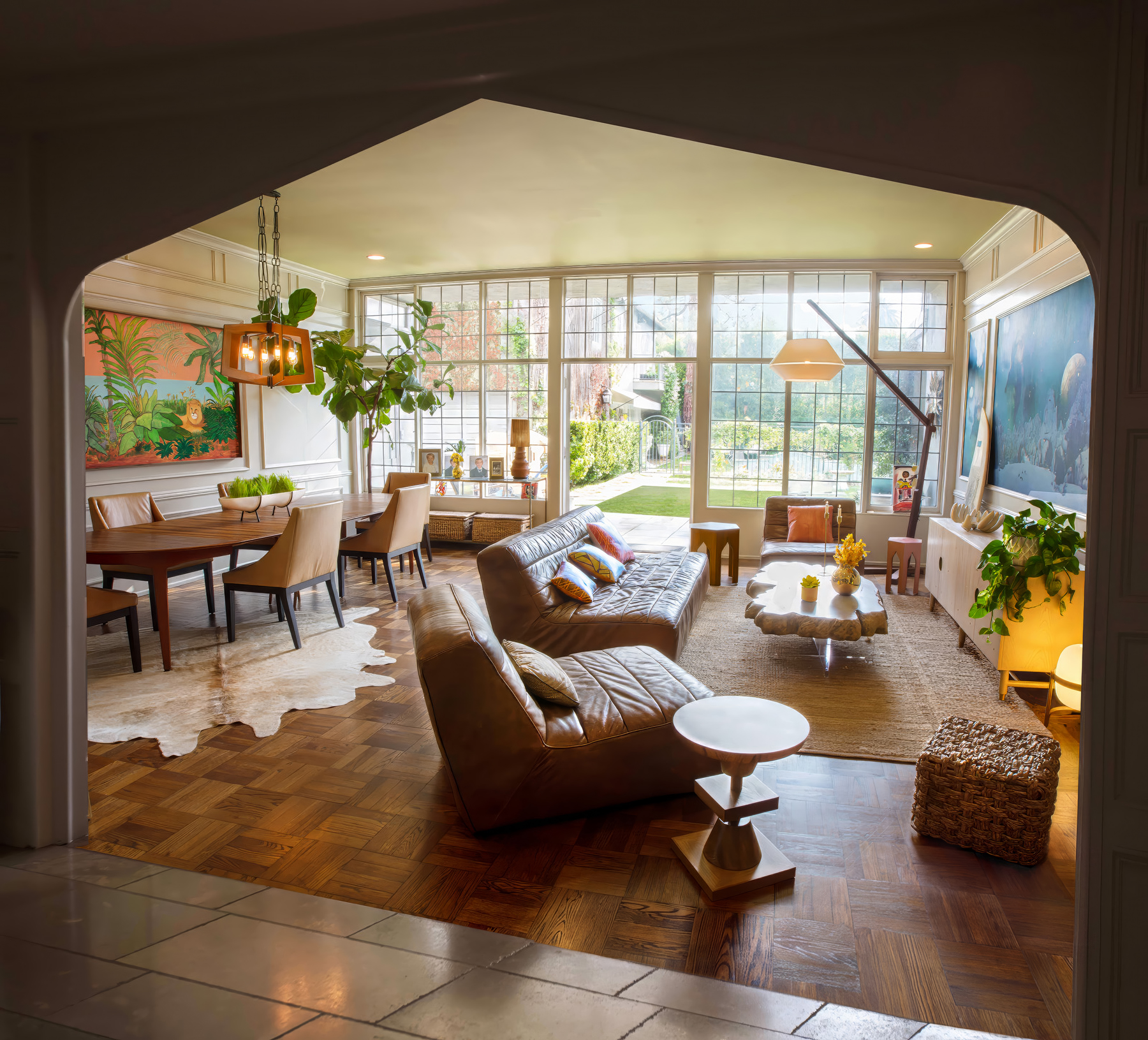 Sunlit living and dining room with leather sectional, parquet floors, and floor-to-ceiling windows opening to a garden, viewed through an arched doorway